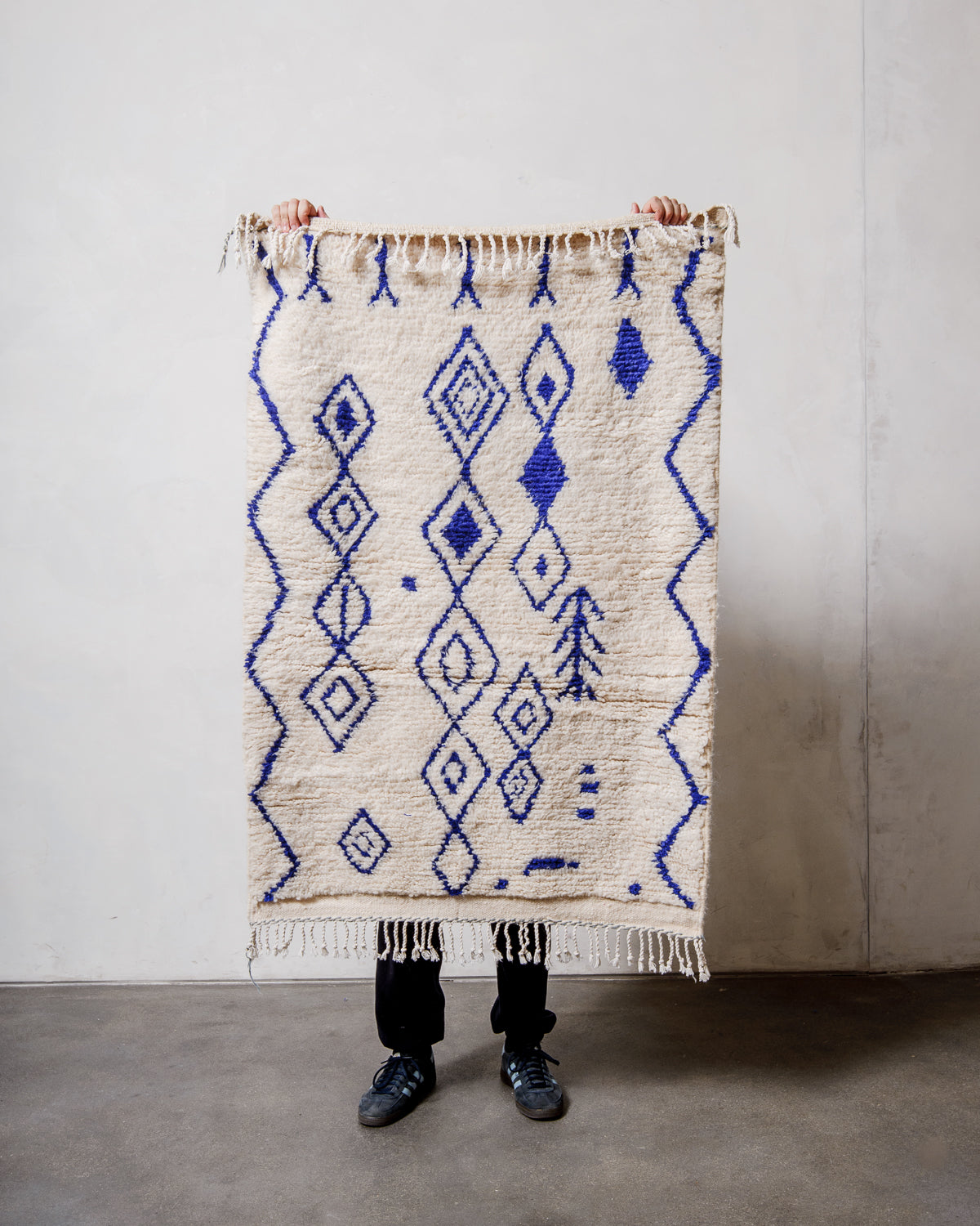 Berberlin Azilal rug in cream wool with blue cotton design being held up in front of a white wall.